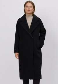 Czarny wełniany płaszcz o długiej, prostej sylwetce, szerokie klapy i boczne kieszenie; posiada pojedyncze zapięcie na guzik i gładką fakturę.