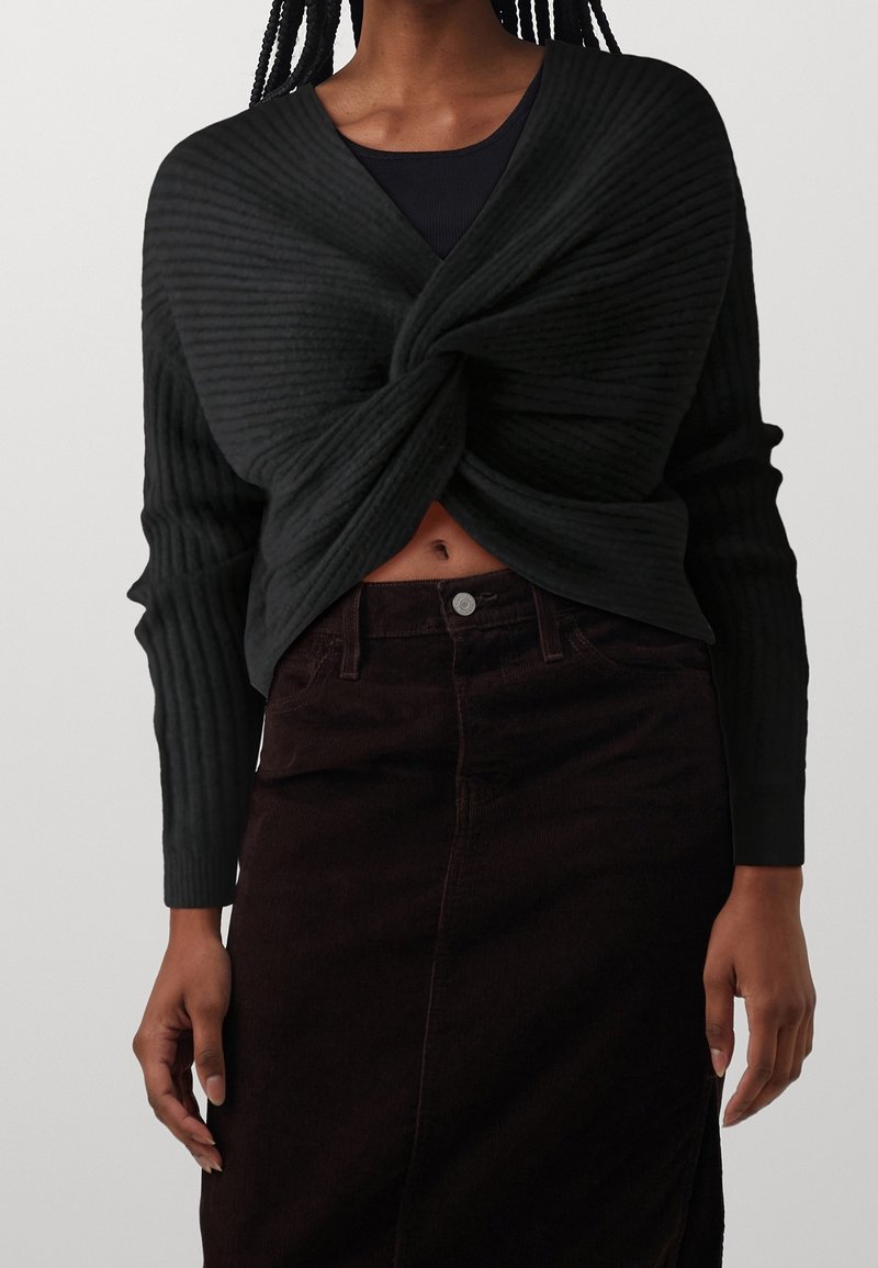 Zwarte gebreide wrap-top met ribbeltextuur, met een gedraaide voorkant, gedragen over een zwarte tanktop en gecombineerd met een donkere corduroy rok.