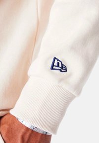 Sudadera color crema con puños acanalados, que presenta un logotipo bordado en azul en la manga. Tejido suave con textura lisa.