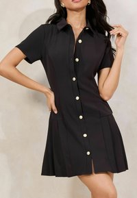 Black short-sleeve shirt dress with a collar, gold buttons, and pleated skirt hem. Smooth fabric with a flattering A-line silhouette.