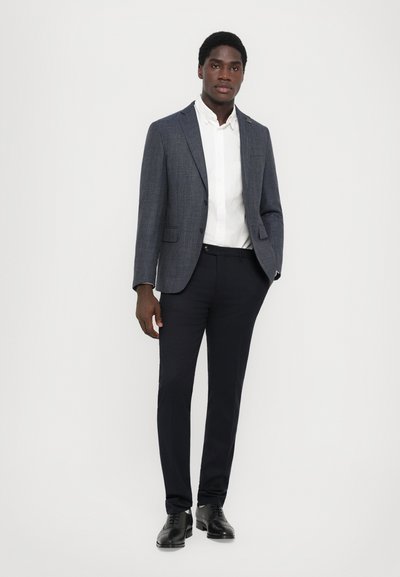 Marineblazer mit karierter Musterung, darunter ein weißes Hemd und schwarze maßgeschneiderte Hosen. Schwarze Dressschuhe vervollständigen das Outfit.