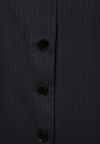 Marineblau gestreuter Stoff zeichnet sich durch schwarze Knöpfe aus; vertikale Streifen erzeugen eine strukturierte Optik, die den Fokus auf die Schneiderdetails und das Handwerk legt.