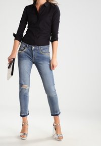 Mujer vestida con una camisa negra de botones, jeans azules rasgados con detalles de perlas, sandalias de tacón alto plateadas, sosteniendo un clutch blanco.