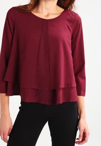 Blouse bordeaux en tissu lisse, avec un col rond, des manches trois-quarts et un design en couches avec des coutures délicates.