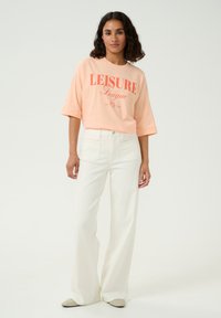 Różowa koszulka z krótkim rękawem z grafiką "LEISURE League" w połączeniu z off-white szerokimi spodniami, które mają gładką teksturę i dopasowany pas.