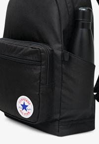 Černý látkový batoh s přední kapsou na zip a boční kapsou držící černou láhev na vodu; má bílý kruhový logotyp jako ozdobu.