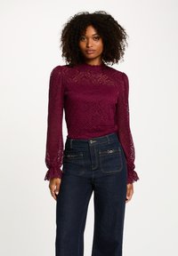 Top in pizzo bordeaux con maniche lunghe, scollatura a onda, motivo floreale, abbinato a jeans in denim scuro a vita alta con dettagli con zip.