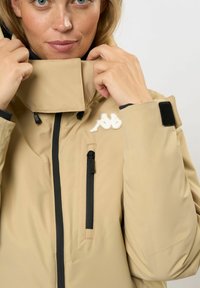 Frau passt den Kragen einer beigefarbenen Jacke mit schwarzen Reißverschlüssen und einem weißen Logo auf der Brust an.