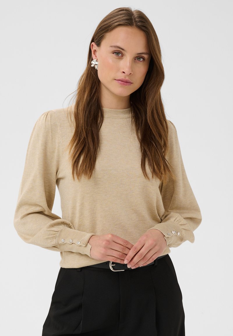 Femme aux longs cheveux bruns portant un pull beige à manches longues avec des boutons aux poignets et un pantalon noir, debout devant un fond clair uni.