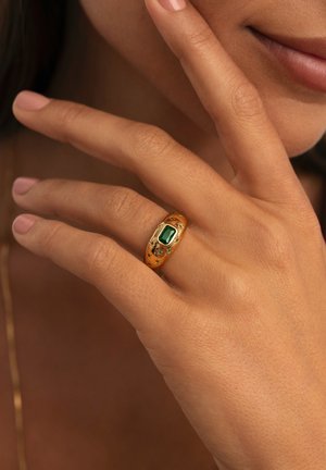 Bague en or avec une pierre précieuse verte rectangulaire et des accents floraux, portée sur une main avec du vernis à ongles nude, montrant une texture de peau lisse.
