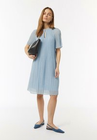 Lichtblauwe geplooide jurk met een sleutelgatnek, met een subtiel patroon. Gecombineerd met een zwarte handtas en blauwe slingback flats.