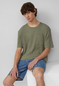 Jasnooliwkowy t-shirt z krótkim rękawem o fakturowanym wzorze waflowym oraz kieszonką na piersi, w połączeniu z niebieskimi szortami dżinsowymi do kolan.