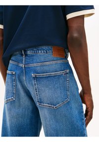 Vue arrière d’une personne portant un jean en denim bleu coupe décontractée et une chemise bleu marine avec des bords de manches blancs.