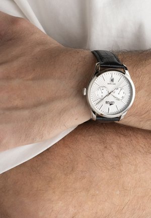 Muñeca con reloj analógico plateado que presenta una esfera texturizada blanca, correa de cuero negro, dos subesferas y ventana de fecha a las 12 en punto.