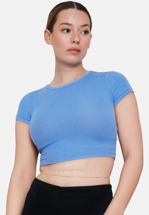 Kobieta z zaczesanymi do tyłu włosami, ubrana w dopasowany, jasnoniebieski krótki t-shirt oraz czarne spodnie z delikatnym łańcuszkowym paskiem w talii.