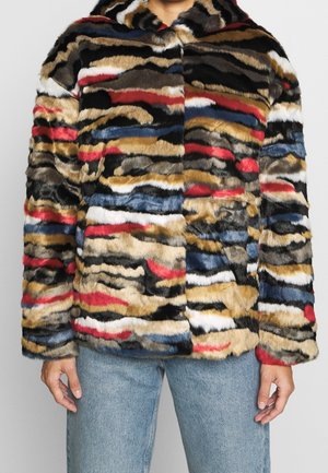 Personne portant une veste en fausse fourrure rayée multicolore avec des tons rouges, bleus, noirs, blancs et beige, accompagnée d'un jean bleu clair.