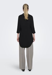 Tunic nera con maniche a tre quarti, caratterizzata da un orlo curvo, abbinata a pantaloni larghi grigi chiari e tacchi chunky neri.