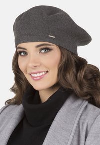 Berretto di maglia grigio con design slouchy e piccolo accento metallico. Indossato con un maglione nero a collo alto e un cappotto grigio.