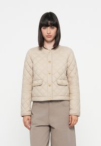 Beige quilted jacket with gold buttons, two front pockets, and a rounded neckline. Soft texture and cropped fit, paired with beige trousers.