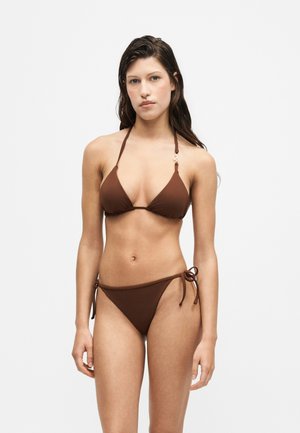Jeune femme debout portant un bikini triangle marron foncé avec des liens à nouer, sur un fond clair uni.