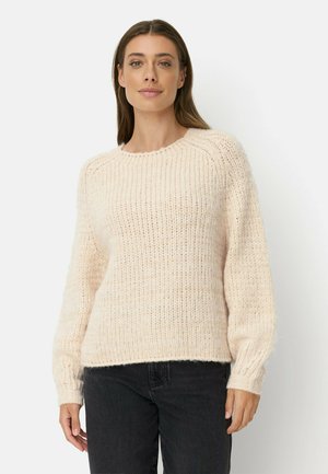 Vrouw met lang bruin haar, gekleed in een crèmekleurige geribbelde gebreide trui en zwarte broek, staand tegen een witte achtergrond.