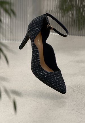 Scarpa nera con tacco alto, con tomaia in tessuto testurizzato, punta affusolata e cinturino alla caviglia con una fibbia dorata. La suola è marrone.