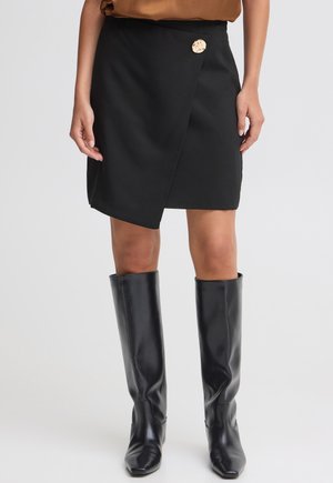 Femme portant une jupe portefeuille noire avec un détail de bouton doré, associée à des bottes en cuir noires montant jusqu'aux genoux, debout devant un fond uni.