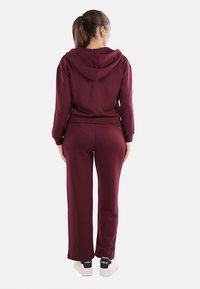 Felpa con cappuccio bordeaux e pantaloni abbinati, realizzati in tessuto morbido; presenta una vestibilità rilassata e polsini elastici; abbinati a sneakers bianche.