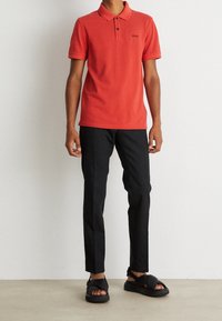 Man som bär en röd kortärmad polotröja, svarta slim-fit byxor och svarta sandaler med korsremmar, stående mot en vit vägg.