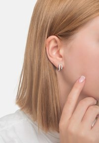 Silver musiknot-formad studörhänge som bärs på örsnibben. Enkelt, minimalistisk design med en glänsande yta som kompletterar ljusa hårfärger.