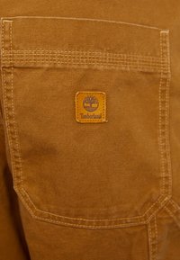 Taschino in tessuto color tan con dettagli cuciti e una toppa quadrata in pelle con un logo di albero e il testo "Timberland".