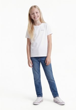 Lächendes Mädchen mit langen blonden Haaren, das ein weißes Calvin Klein T-Shirt, blaue Jeans und weiße Turnschuhe trägt und vor einem weißen Hintergrund steht.
