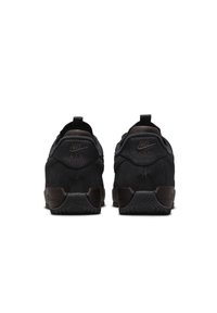 Mustat mokkajalkineet, joissa on ruskeita yksityiskohtia. Mallissa on pyöreä muotoilu, teksturoitu kumipohja ja vetotuki kantapäällä. Nike-logo on kirjailtu taakse.