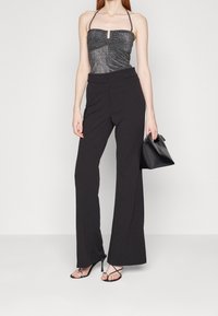 Kobieta ubrana w błyszczący czarny top na szyję, czarne spodnie o wysokim stanie z szerokimi nogawkami, czarne sandały na szpilkach oraz trzymająca czarną torebkę.