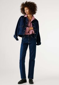 Τζιν μπουφάν σε σκούρο μπλε με χρυσές λεπτομέρειες, σε συνδυασμό με πουκάμισο με κουμπιά κόκκινο και λευκό με ρίγες. Στενά τζιν σε σκούρο μπλε και μαύρα παπούτσια.