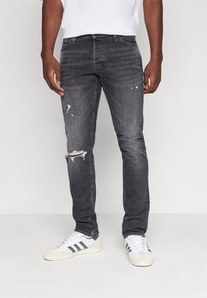 Dark gray jeans with a slim fit, featuring noticeable rips at the knees and faded texture. Worn with white sneakers and a plain t-shirt.