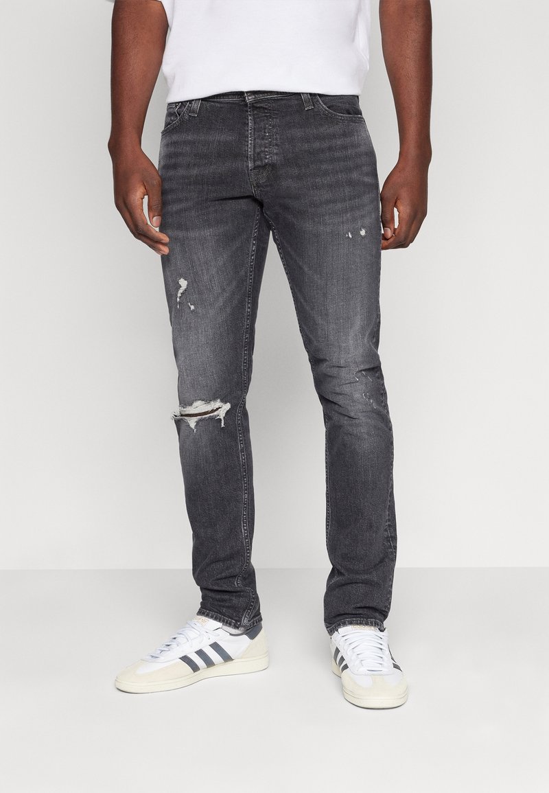 Mörkgrå jeans med en slim fit, med märkbara rivningar vid knäna och urblekt struktur. Bärs med vita sneakers och en enkel t-shirt.