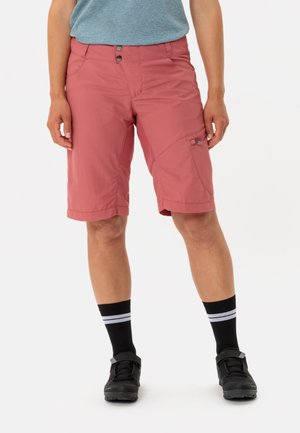Rosa Shorts aus leichtem Material, ausgestattet mit zwei Vordertaschen und einer Seitentasche. Kombiniert mit schwarzen Socken mit weißen Streifen und Schuhen.