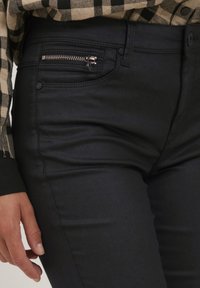Crne traperice imaju zipzarski džep s metalnim detaljima, glatku teksturu i minimalne šavove, u kombinaciji s kariranim flanel košuljom.