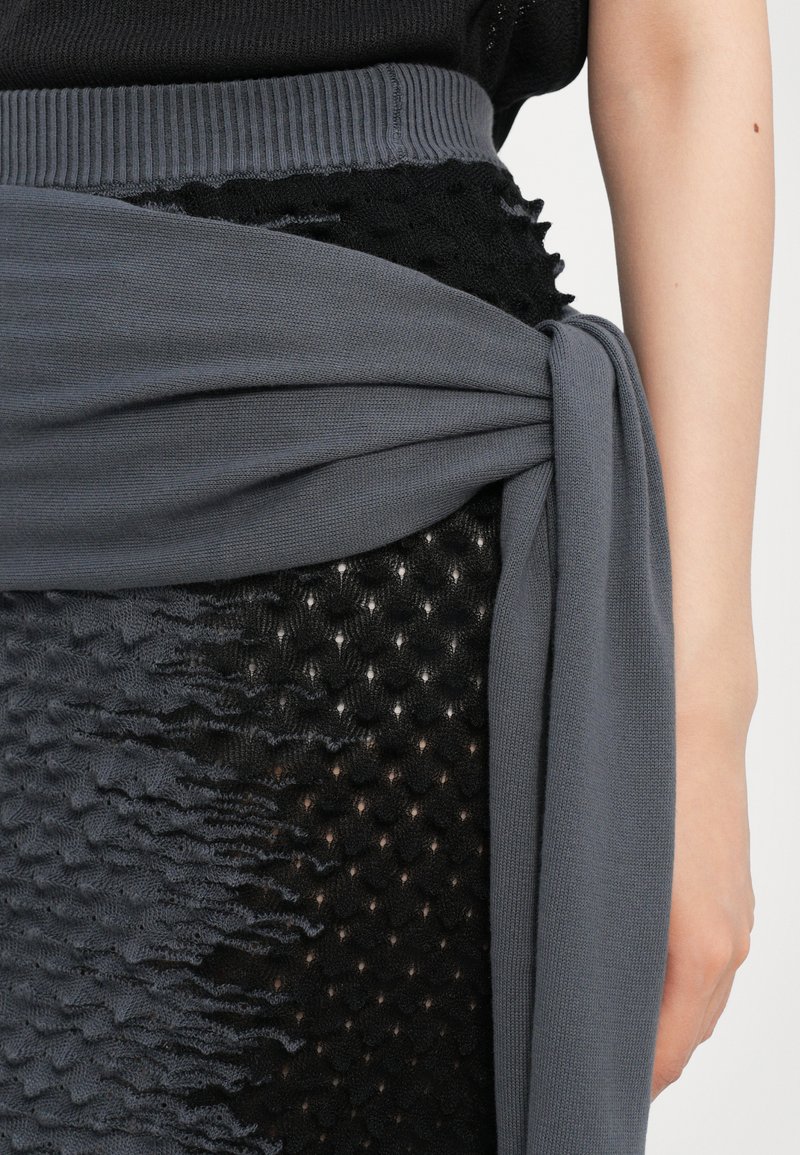 Tessuto a maglia grigia avvolto attorno a una gonna nera, semitrasparente e testurizzata, caratterizzata da un design a strati con dettagli arricciati su un lato.