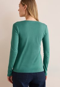 Top a maniche lunghe di colore teal realizzato in tessuto morbido, con scollatura rotonda e forma aderente. Abbinato a jeans scuri per creare contrasto.
