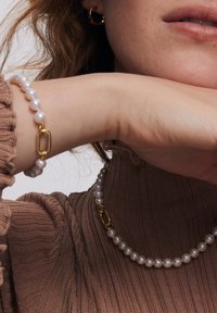 Perłowa bransoletka z akcentem złota i naszyjnik perłowy. Oba mają okrągłe perły, umieszczone na tle fakturowanego brązowego swetra dzianinowego.