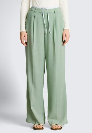 Pantaloni verde chiaro larghi con pieghe frontali e coulisse bianca, indossati con sandali crema e top a maniche lunghe bianco a coste.