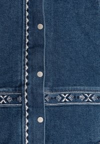 Džínová košile v tmavě modré barvě se stříbrnými kovovými knoflíky, dekorativním bílým a černým vyšíváním a texturovaným povrchem.