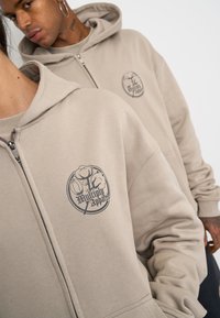 Las sudaderas con cremallera en color beige presentan un logotipo circular con texto y dos diseños florales. Están fabricadas con una tela suave y tienen un corte relajado.