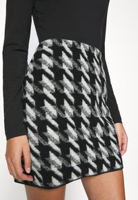 Femme portant une mini-jupe à motif pied-de-poule noire et blanche en tissu duveteux, avec un haut noir à manches longues, se tenant devant un fond uni.