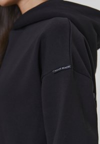Maglione nero con cappuccio in morbido tessuto. Presenta un'etichetta sulla manica sinistra con la scritta "SOUTH BEACH." Inquadratura ravvicinata sul design della manica.