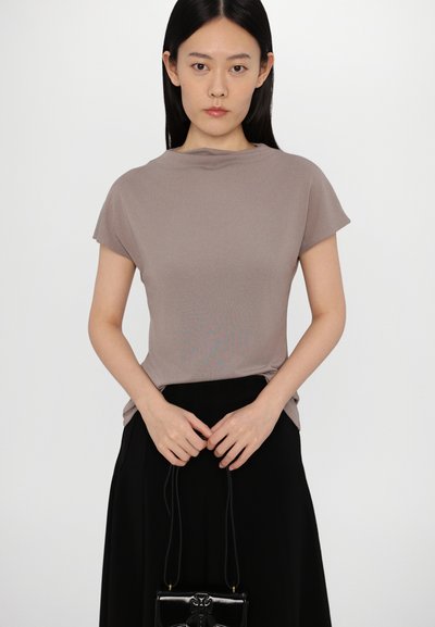 Top a maniche corte in maglia color taupe con scollatura a girocollo discreta, abbinato a una gonna nera e una piccola borsa nera con accessori decorativi.