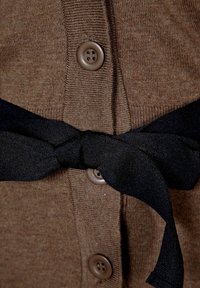 Brauner Cardigan mit geripptem Muster, sechs hellbraunen Knöpfen und einem gebundenen schwarzen Stoffgürtel in der Taille.