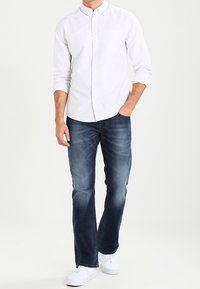 Ljust grå långärmad skjorta med ståkrage och liten prickmönster, kombinerad med mörkblå denimjeans och vita sneakers.